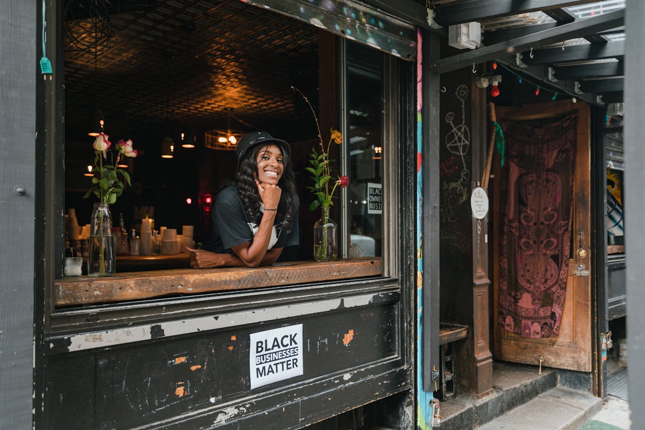 A cheerful businesswoman leaning out of her café window, showcasing a black-owned business with vibrant flowers and decor.