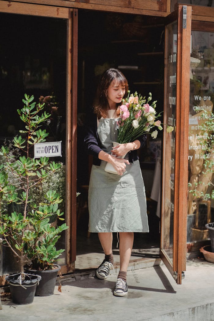 Asian female florist standing at shop entrance with a bouquet of flowers, smiling under sunlight.